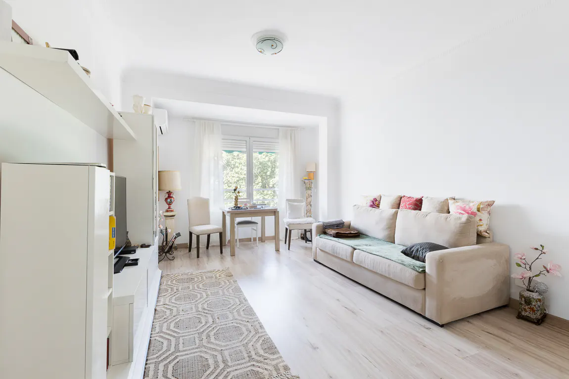Bright living room with white walls, light wood floors, and a beige sofa with colorful pillows. A small dining table sits near the window.