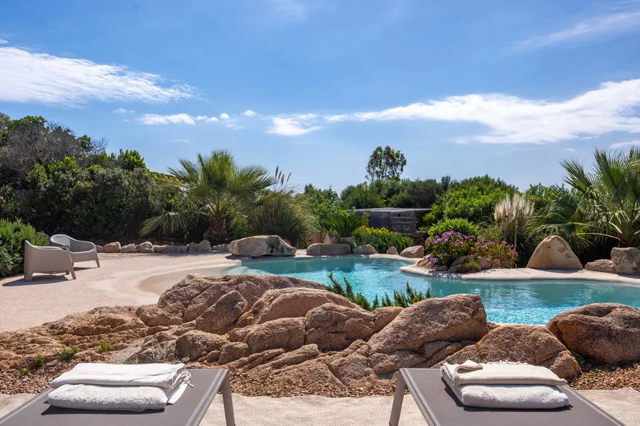 Luxury pool area with lounge chairs, towels, rocks, and lush greenery under a blue sky.