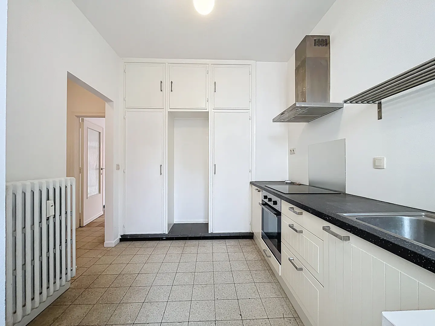 A bright kitchen with white cabinets, black countertops, and a stainless steel range hood. A radiator is on the left.