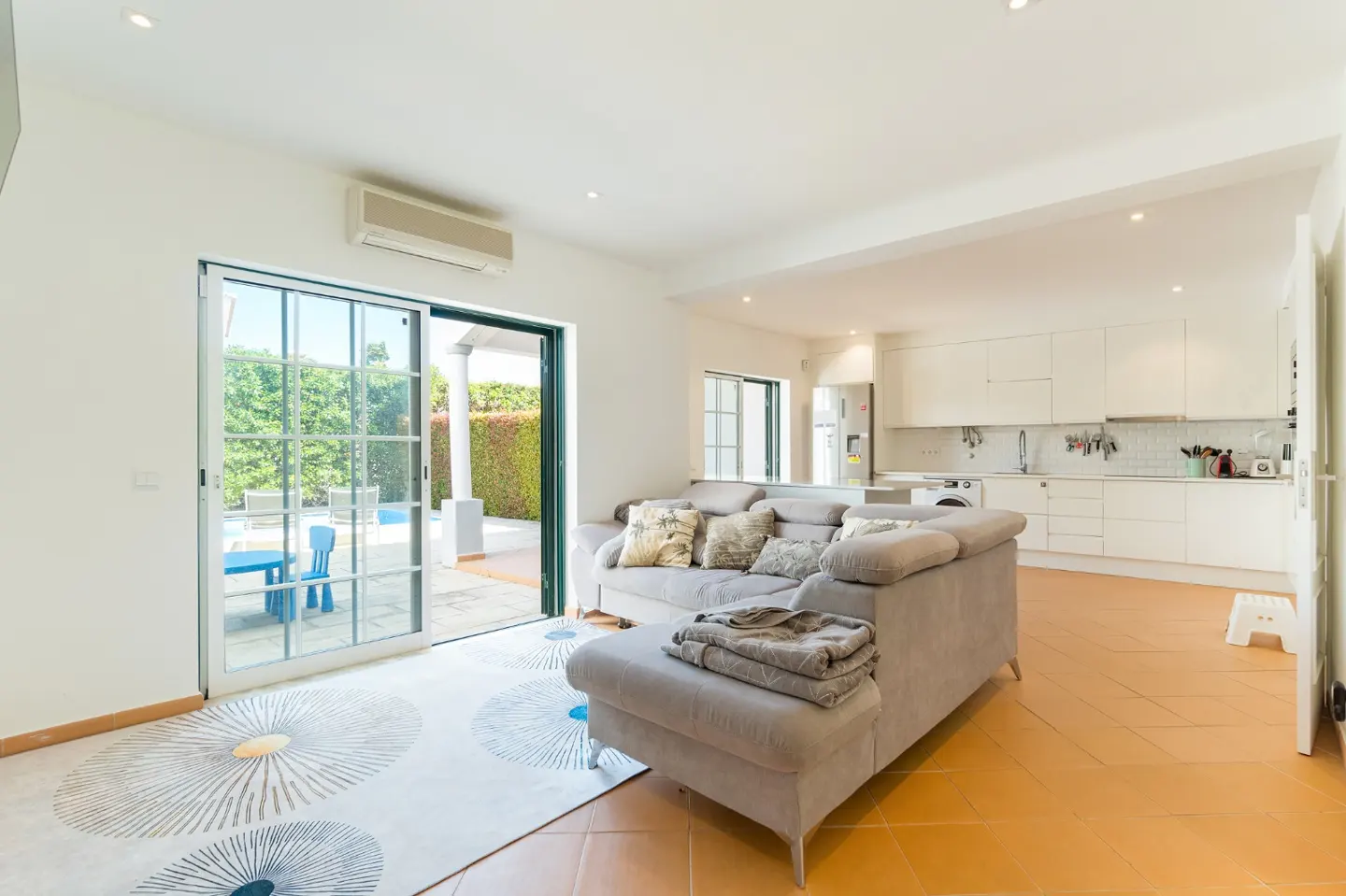 Bright, open-plan living room with a gray sofa, white walls, and terracotta tile floors. Sliding glass doors lead to a patio with a blue table and chairs. A white kitchen is visible in the background.