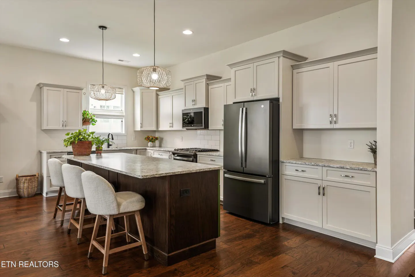 A modern kitchen with white cabinets, a dark island with a marble countertop, and three stools. A black refrigerator is on the right.