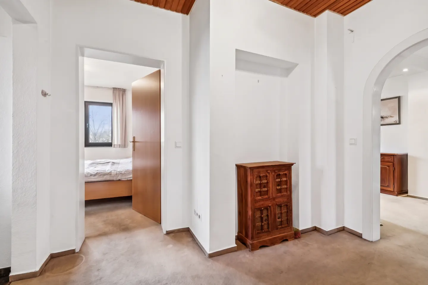 Interior view of a hallway with white walls, a wooden ceiling, and a brown cabinet. A bedroom is visible through an open door.