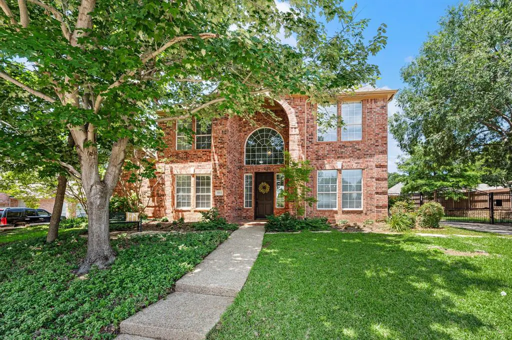 Two-story red brick house with an arched entryway, a concrete walkway, and a green lawn under a large shade tree.
