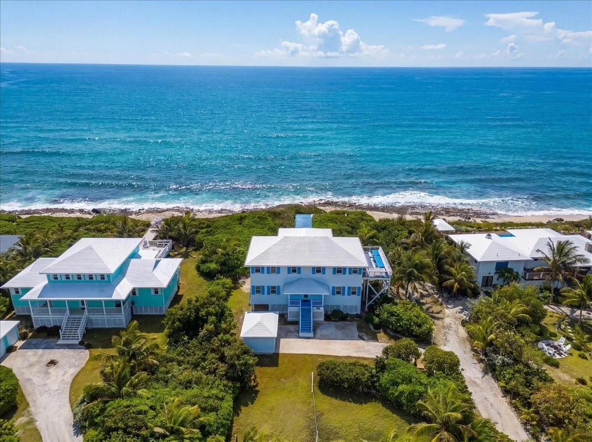 Aerial view of three light blue beach houses with white roofs, lush green vegetation, and turquoise ocean backdrop.
