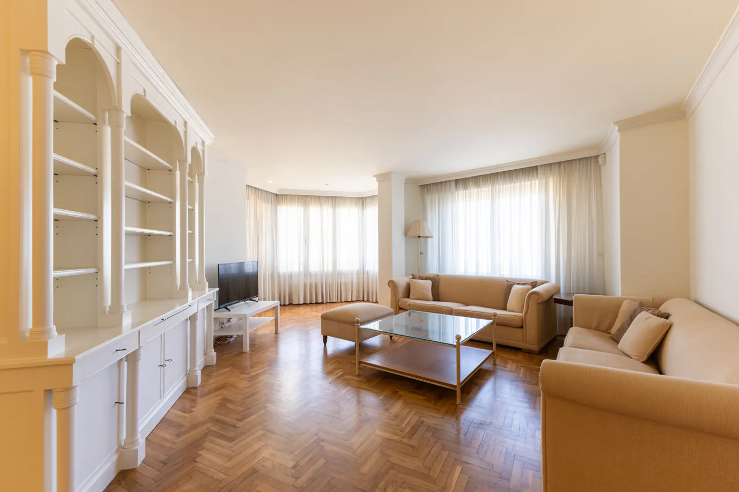 Bright living room with herringbone wood floors, beige sofas, glass coffee table, and a large white built-in bookshelf.