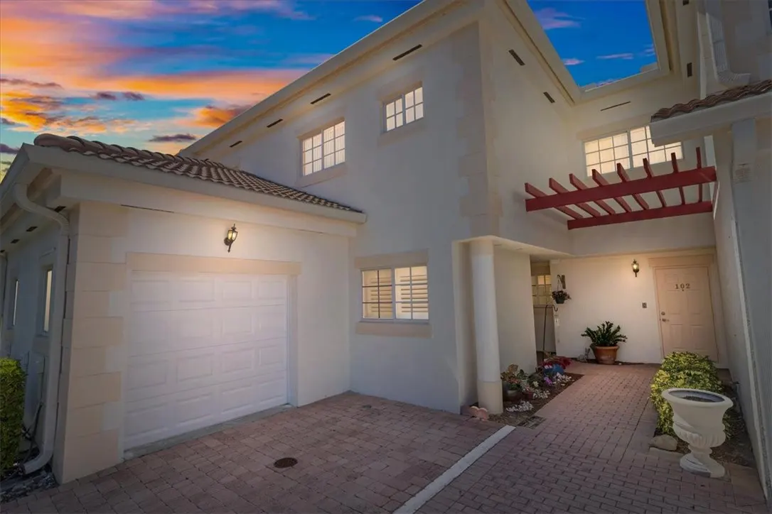 Exterior of a two-story white townhouse with a garage and brick walkway at sunset. Red pergola above the front door.