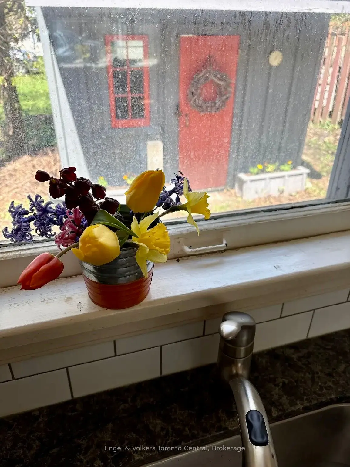 A kitchen window with a flower arrangement in front of a shed with a red door.