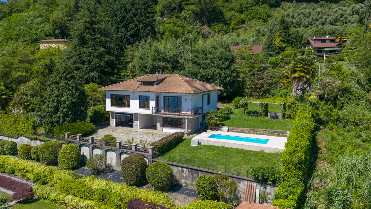 Aerial view of a white house with a brown roof, a pool, and lush green landscaping.