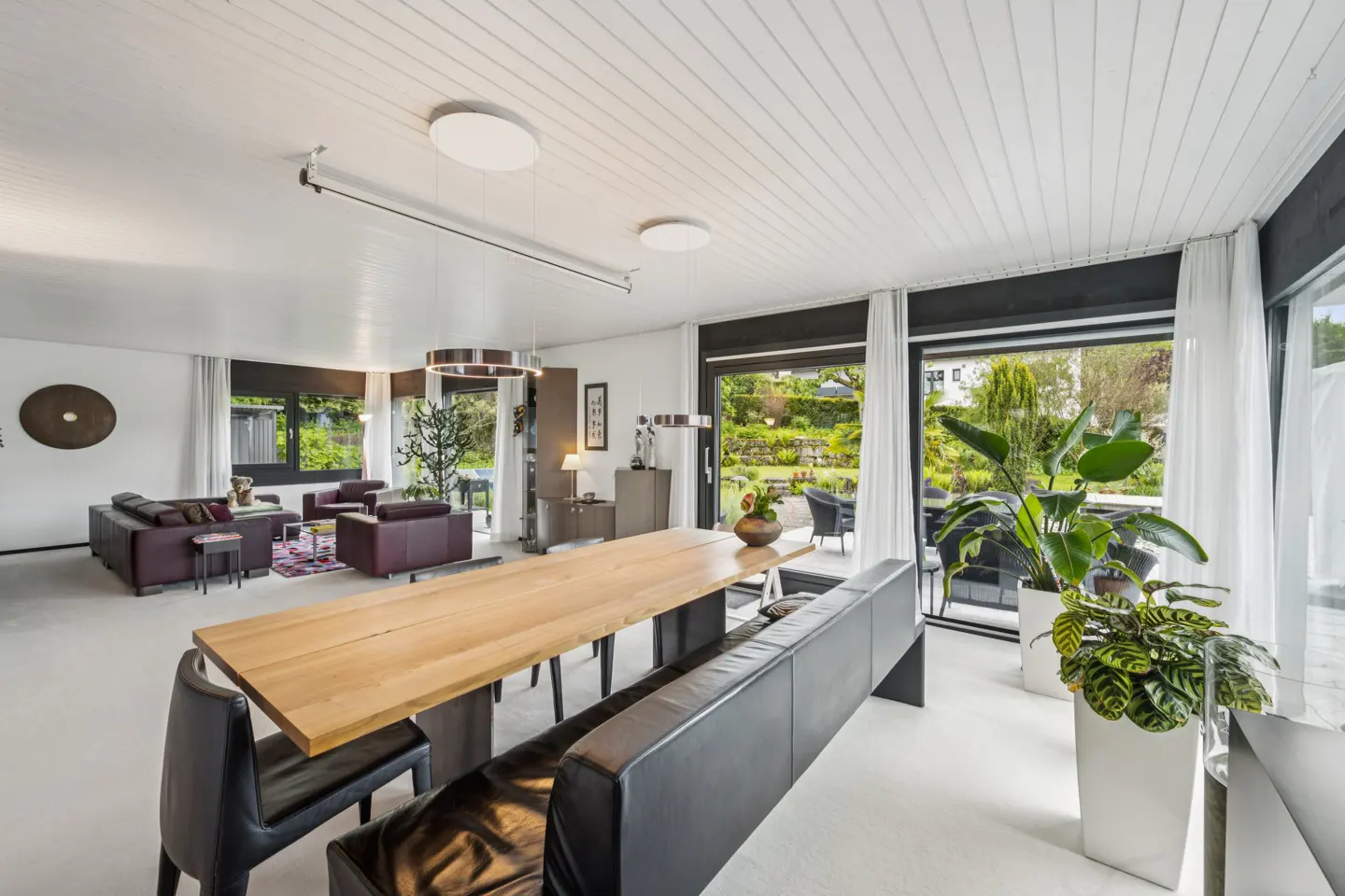 Modern open-plan living area with a long wooden dining table, black leather bench, and a view of a lush green garden.