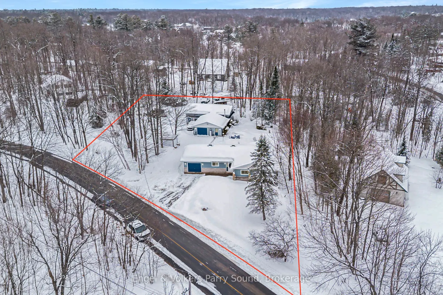Aerial view of a blue house surrounded by snow-covered trees, outlined in red, with a road and car in front.