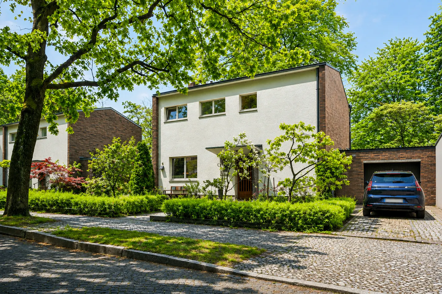 Modern white house with brick accents, green trees, and a blue car parked in the garage. Cobblestone driveway and manicured hedges.