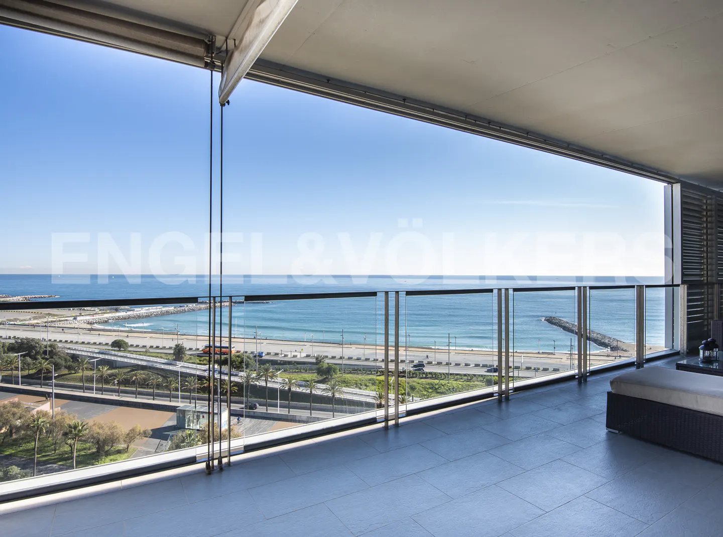 Balcony view of the ocean with a glass railing, a highway, and a sandy beach under a clear blue sky.