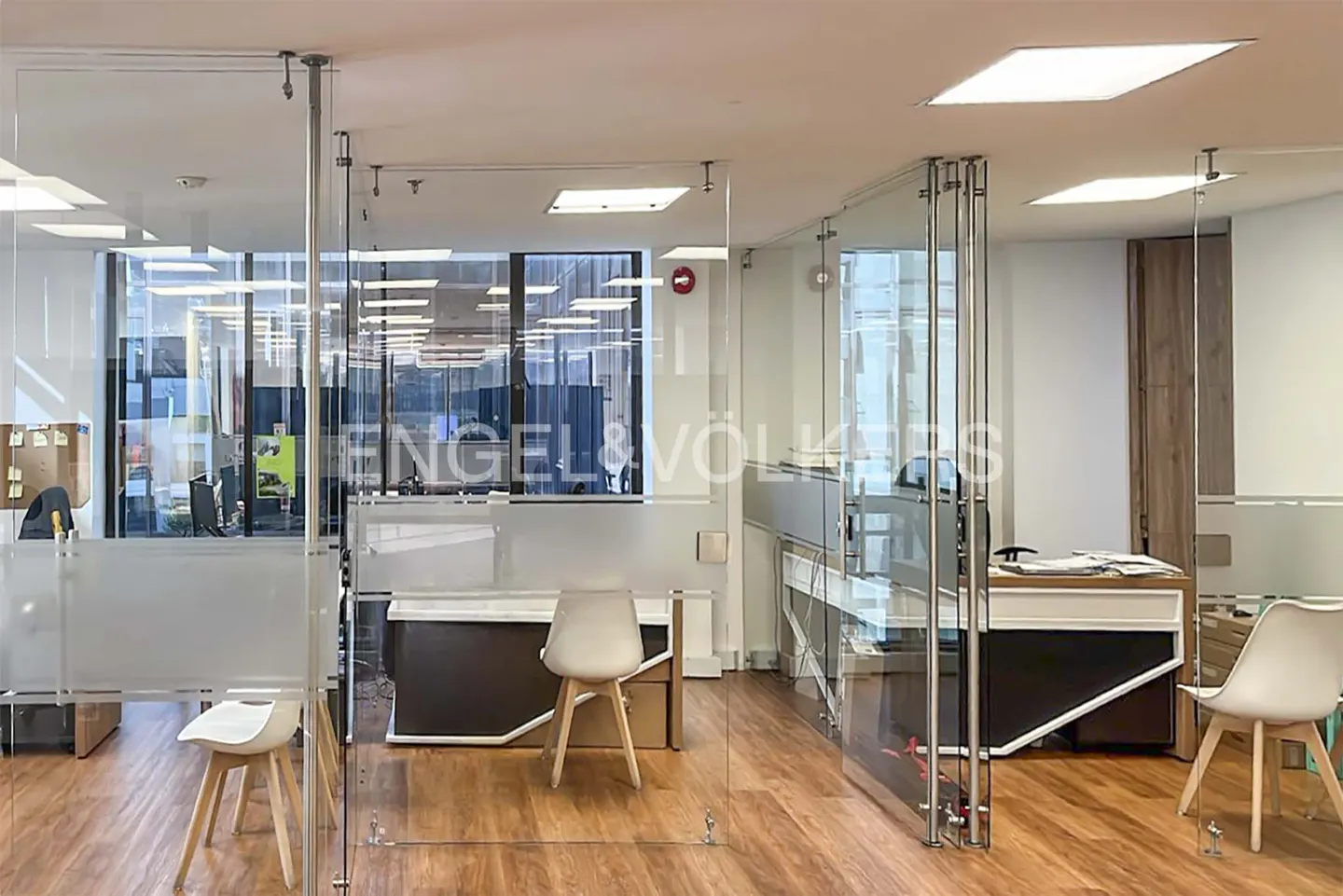 Office interior with glass walls, wood floors, and white chairs. The Engel & Völkers logo is visible on the glass.