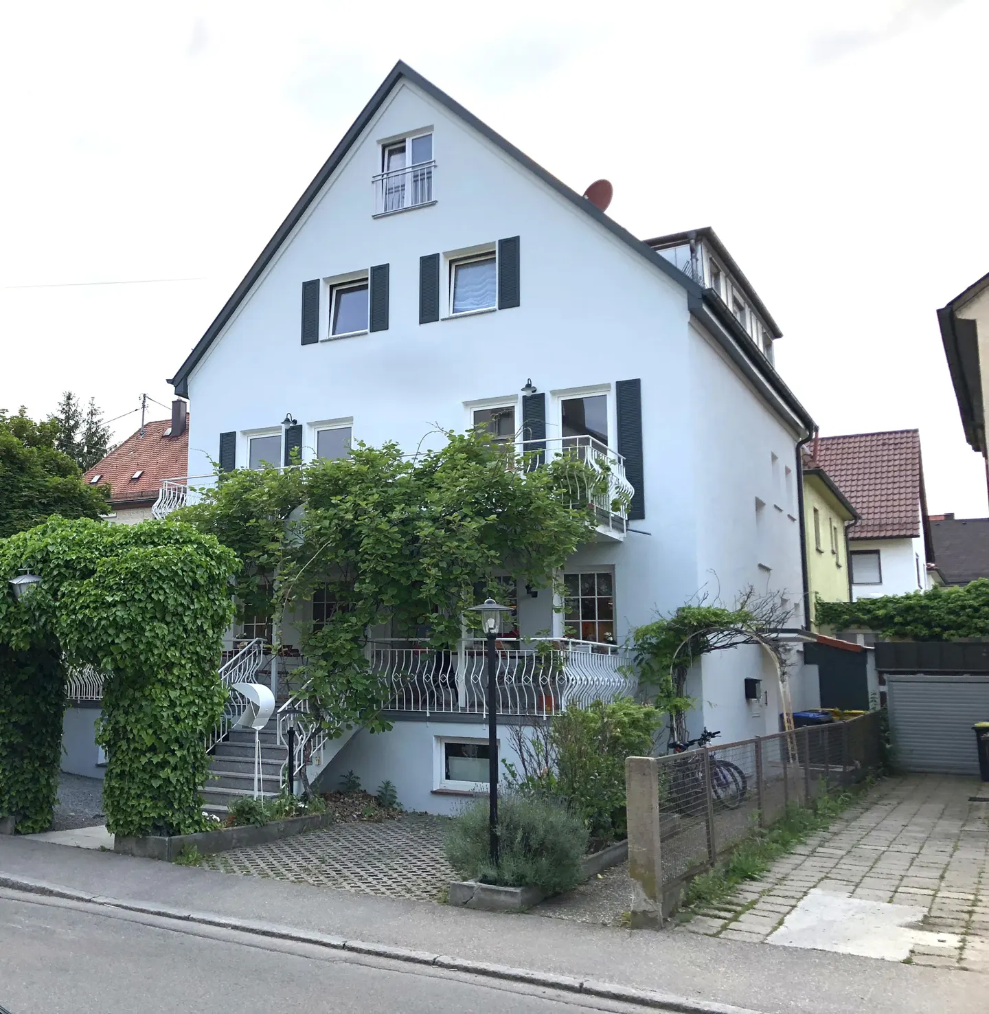 Two-story light blue house with dark shutters and a black roof. Green vines cover the entrance archway and balcony. A paved driveway leads to a garage.