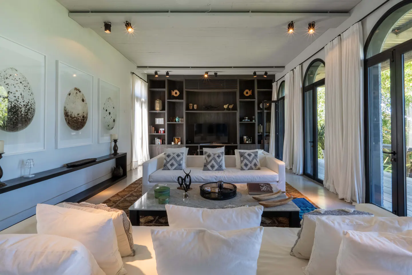 Bright living room with white sofas, pillows, and walls. Large egg art hangs above a dark console. Built-in shelves surround a TV. Arched windows with white curtains.