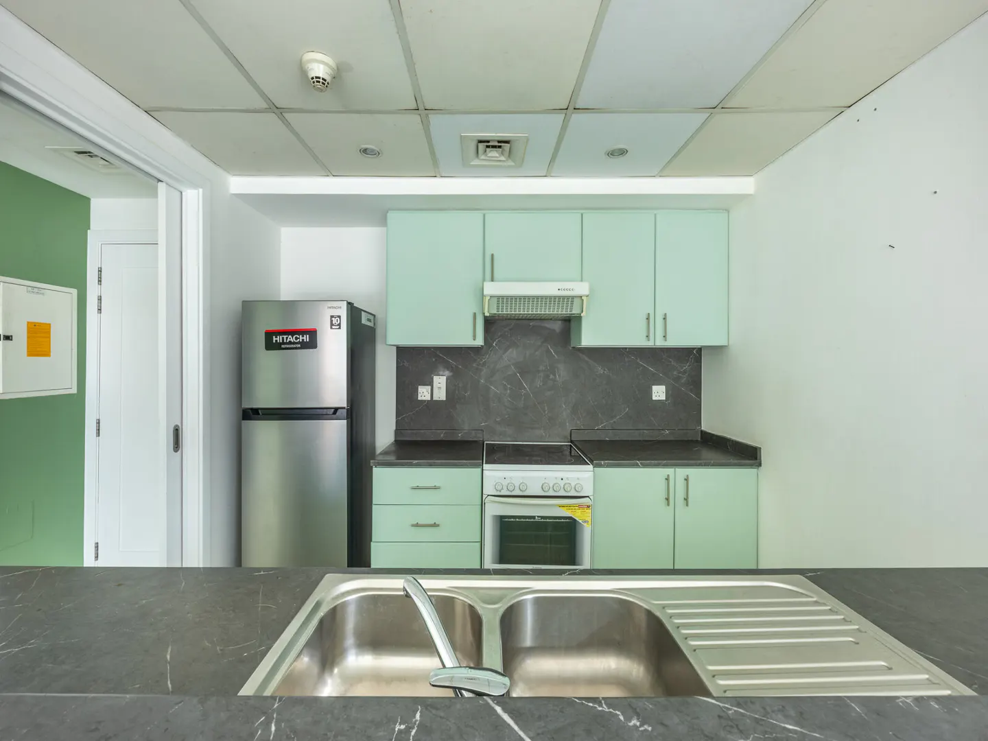A kitchen with light green cabinets, a stainless steel fridge, and a double sink with a black countertop.
