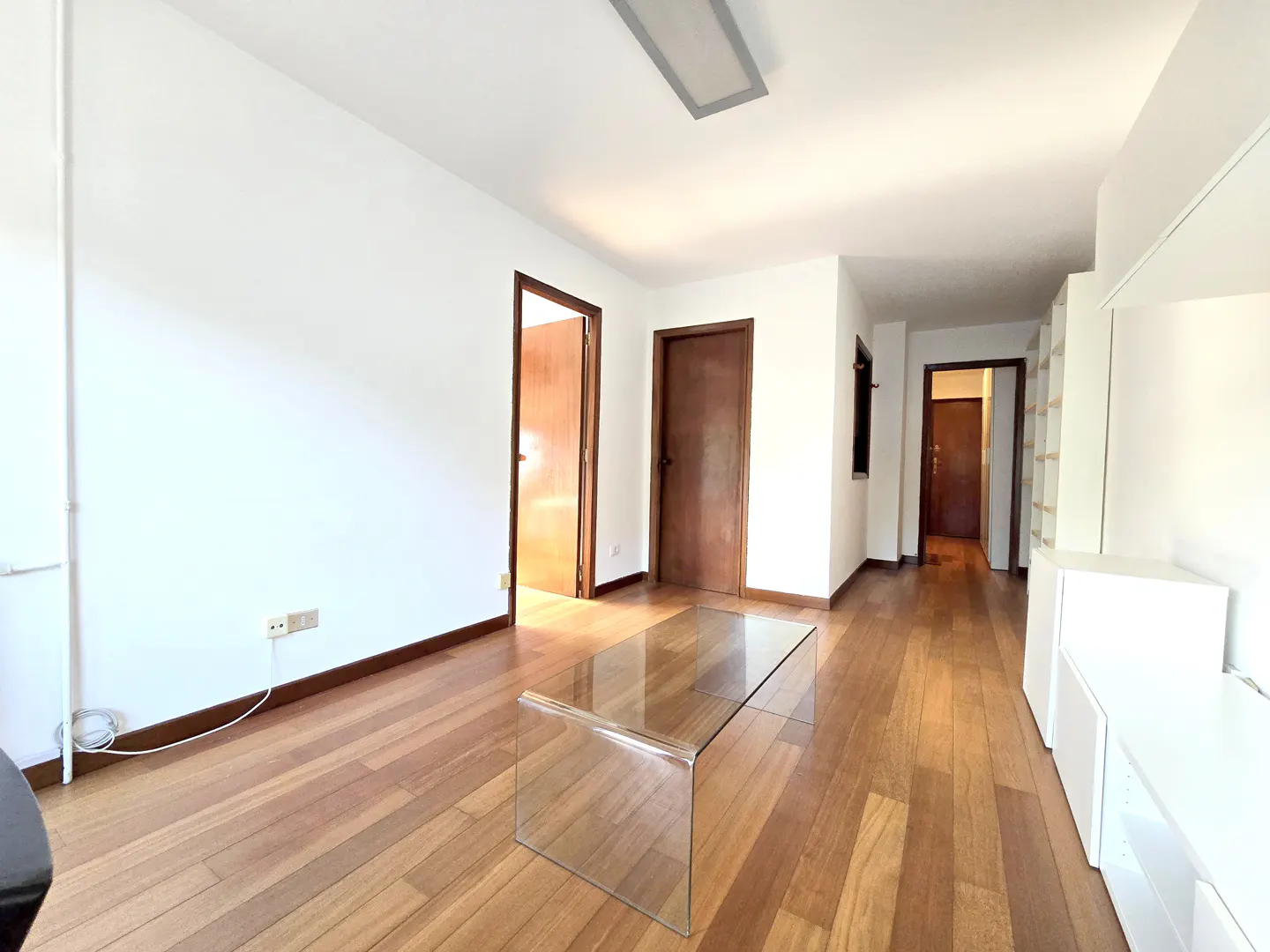 Bright, empty living room with hardwood floors, white walls, and a glass coffee table. Two brown doors and white built-in shelves are visible.