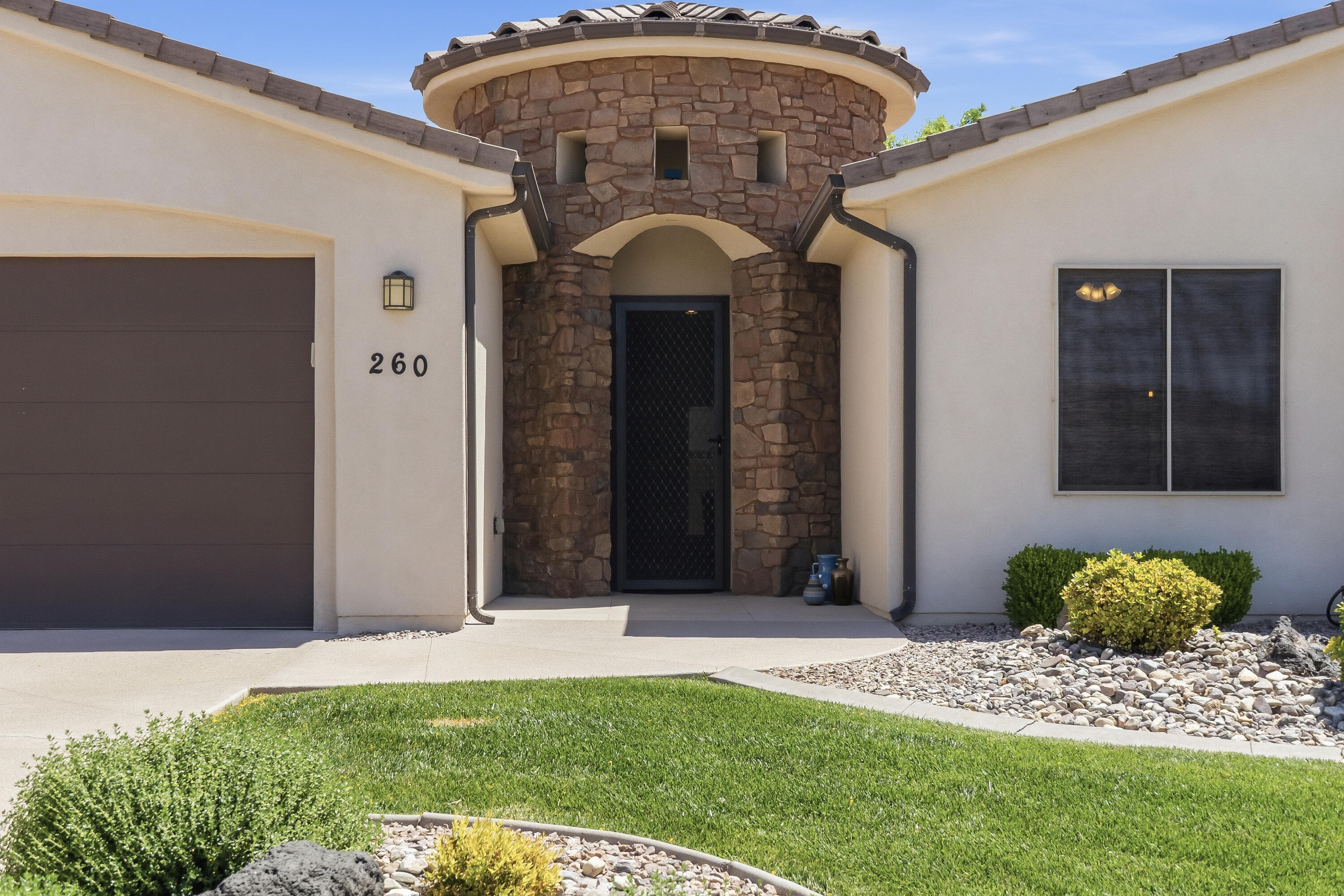 Front exterior of a beige stucco house with a stone turret entrance, dark garage door, and green lawn.
