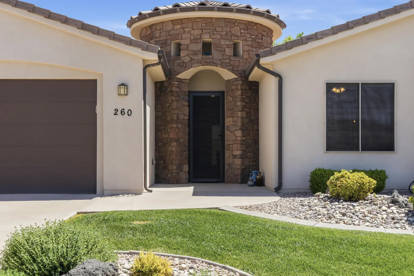Front exterior of a beige stucco house with a stone turret entrance, dark garage door, and green lawn.