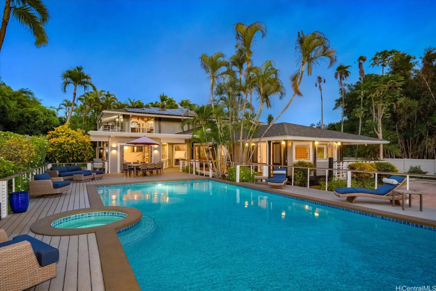 A backyard with a pool, hot tub, deck, and a house with solar panels at dusk.