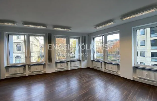 Empty office space with dark wood floors, white walls, and large windows showing city views. Radiators are under the windows. Engel & Völkers logo is visible.