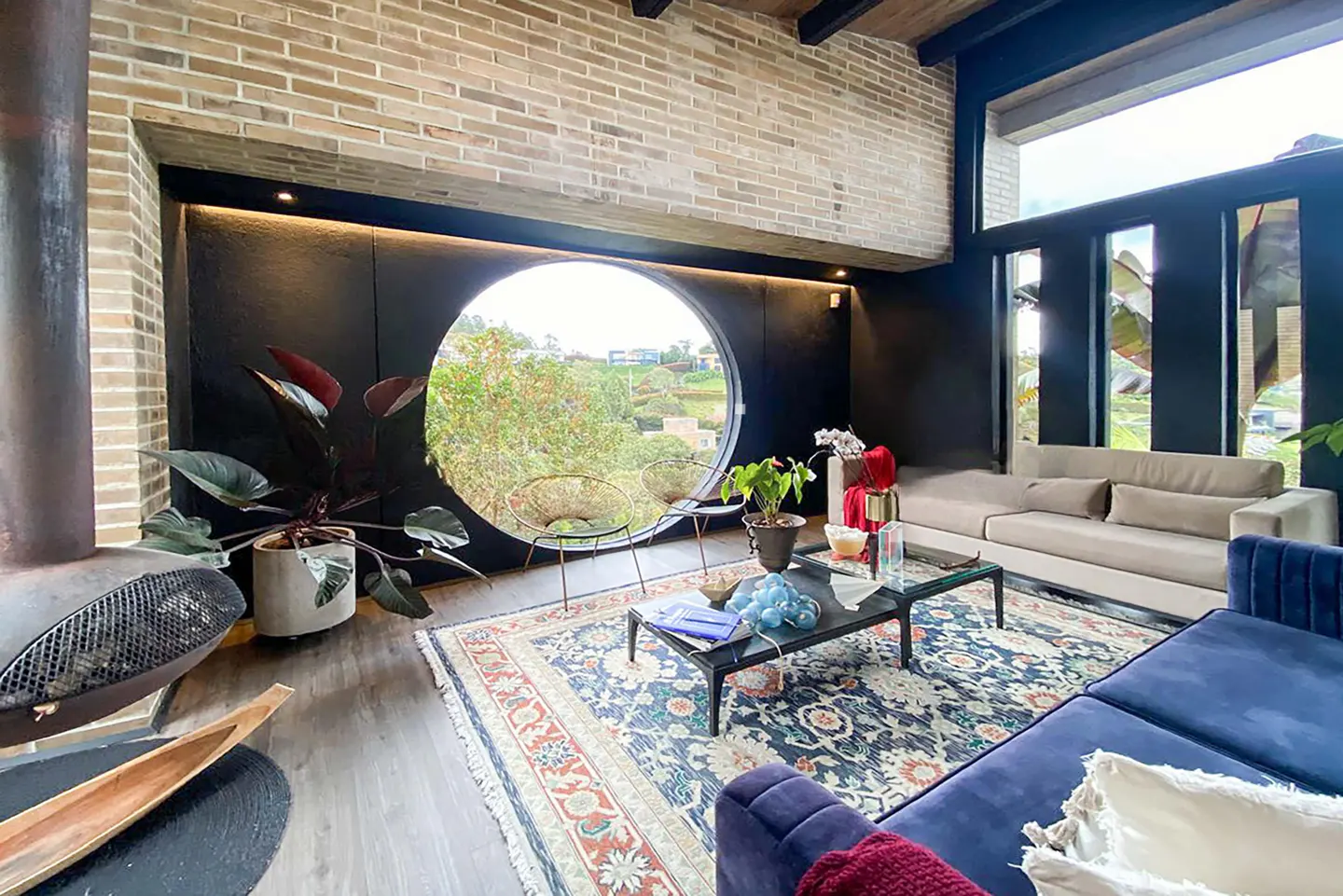 Living room with brick walls, a round window, and a blue patterned rug. Sofas are beige and blue. Plants are placed around the room.