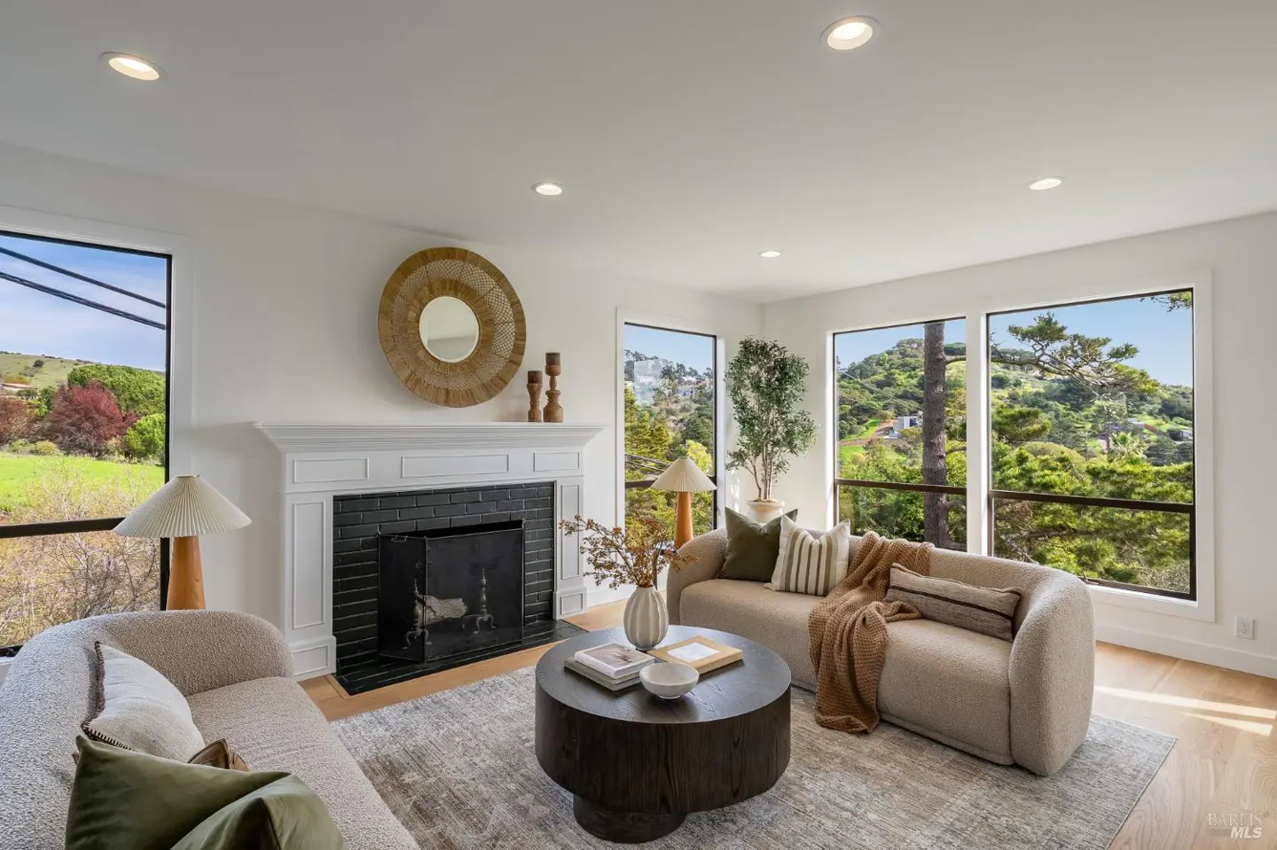 Bright living room with two beige sofas, a round coffee table, and a black fireplace. Large windows show a green, hilly landscape.