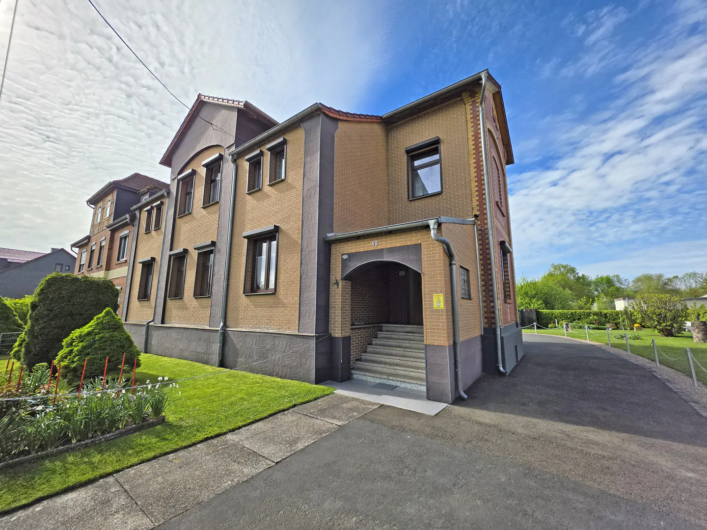 Two-story tan brick building with dark trim, arched entryway, concrete steps, and a paved driveway under a blue sky.