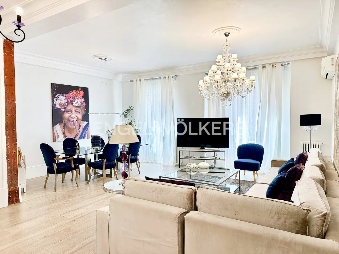 Bright living room with a beige sectional, blue velvet chairs, a glass dining table, and a large crystal chandelier.