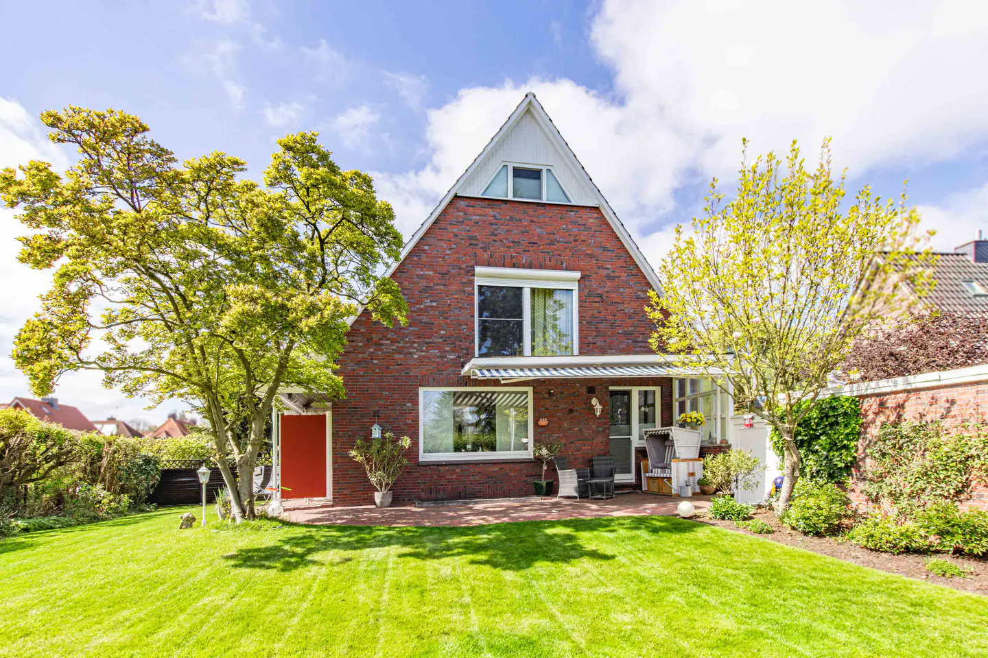 Two-story brick house with a triangular roof, green lawn, trees, and a patio with chairs.