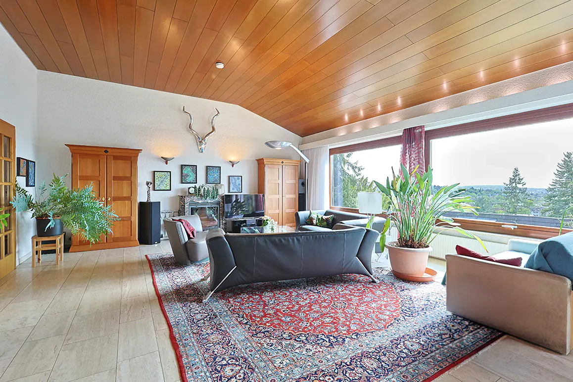 Spacious living room with wood-paneled ceiling, large red patterned rug, leather sofas, and a scenic window view.
