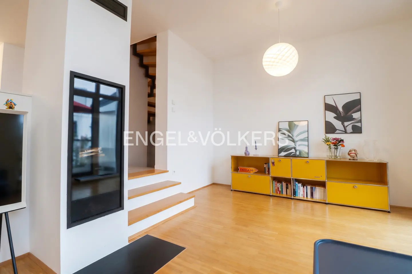 Bright living room with hardwood floors, white walls, and yellow storage. A modern fireplace and open staircase add architectural interest.