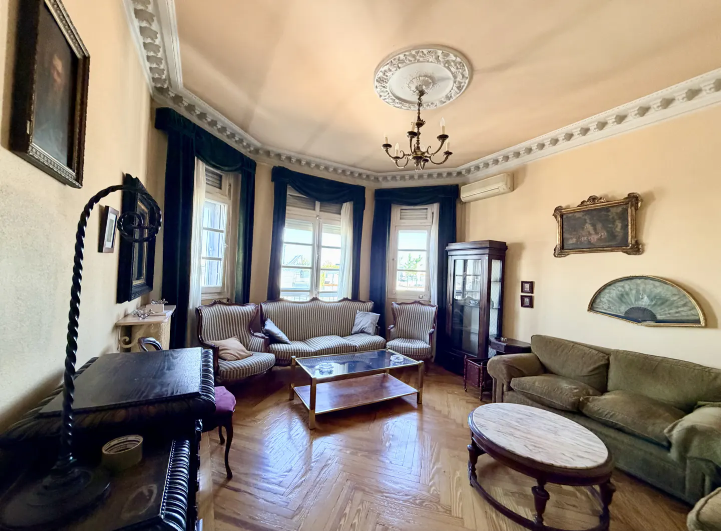A living room with striped sofas, a glass coffee table, and herringbone wood floors. The walls are peach, and there are dark green curtains.