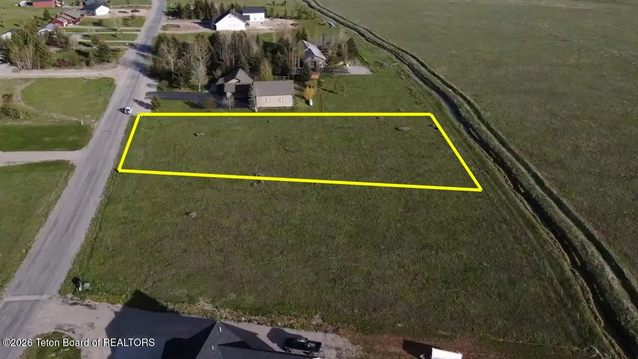 Aerial view of a vacant, grassy residential lot outlined in yellow, situated next to a road and neighboring houses.
