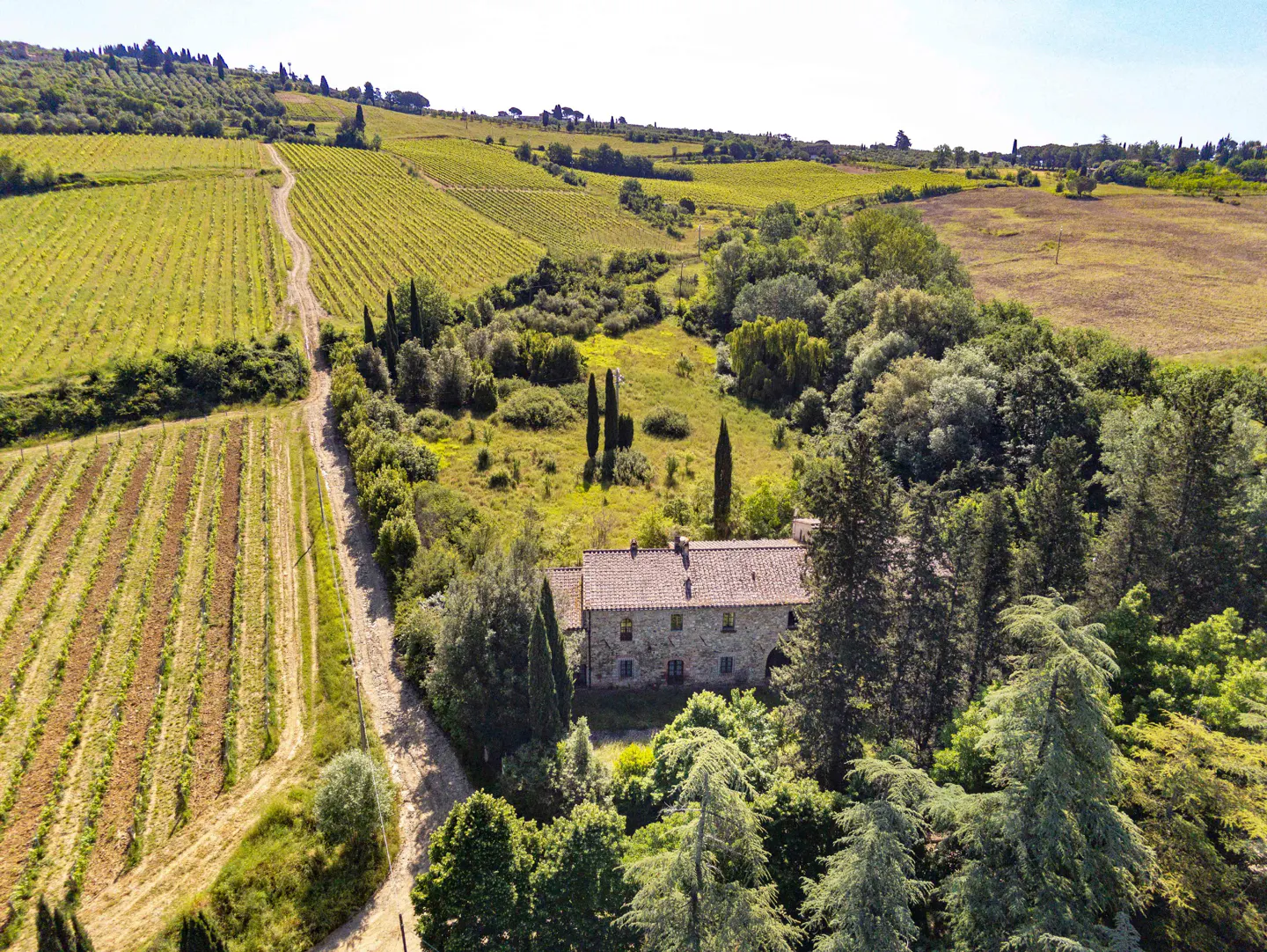 Aerial view of a stone farmhouse with a red tile roof, surrounded by green trees and vineyards in the countryside.