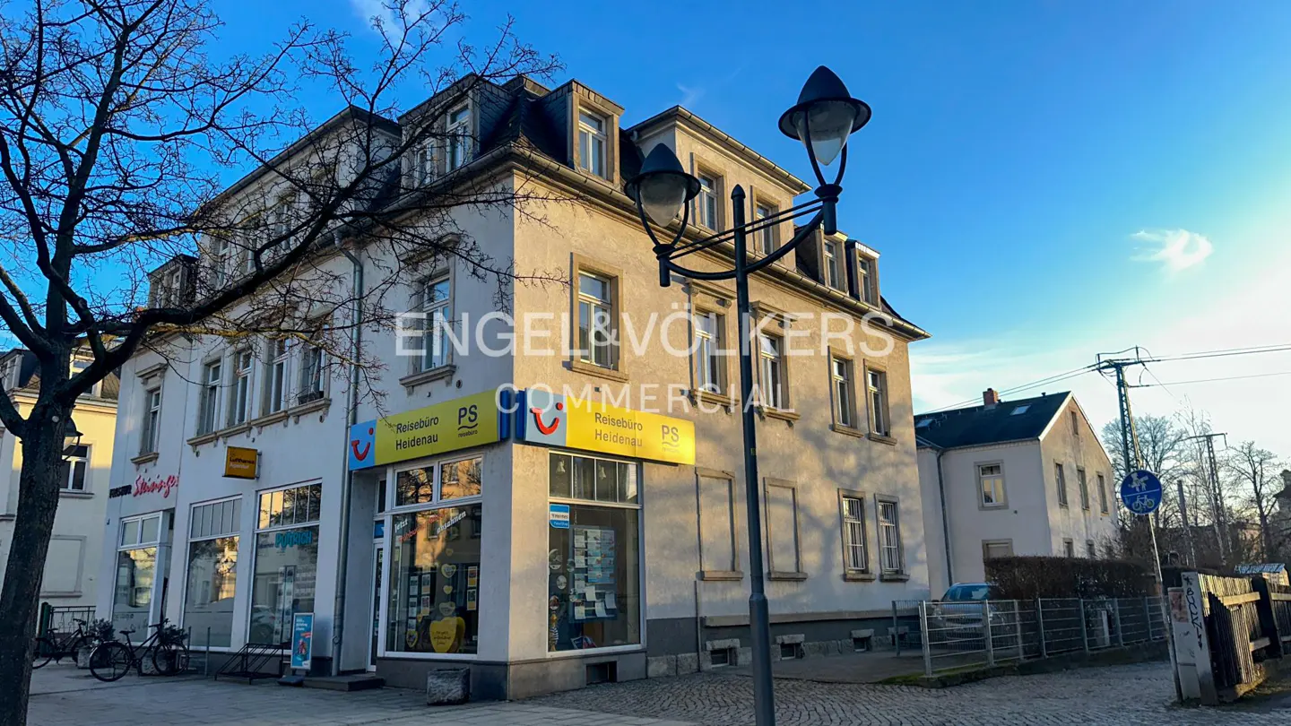 Exterior view of a three-story commercial building with Engel & Völkers signage, travel agency, street lamp, and blue sky.