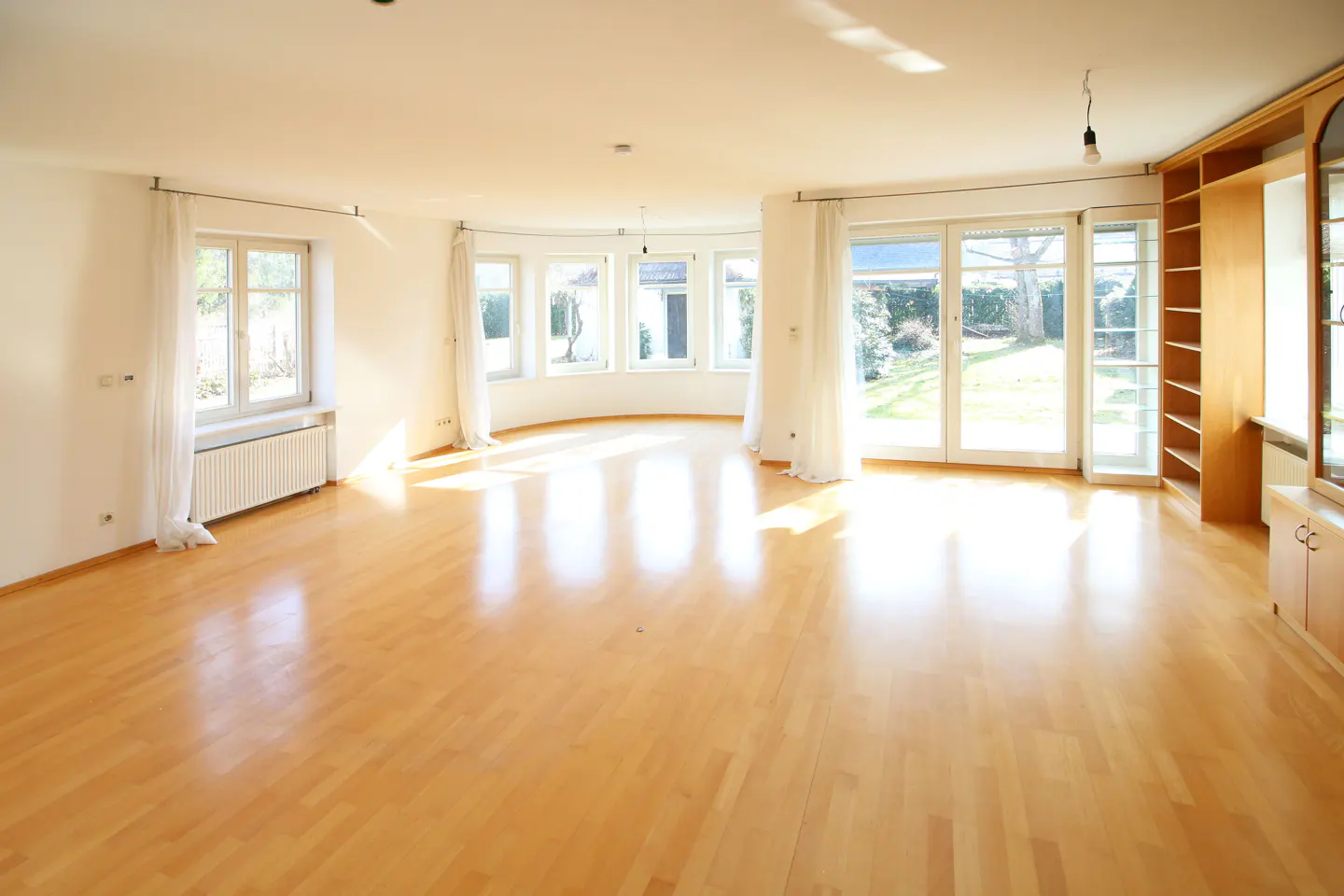 Bright, empty living room with light wood floors, white walls, and multiple windows. A built-in bookcase is on the right.