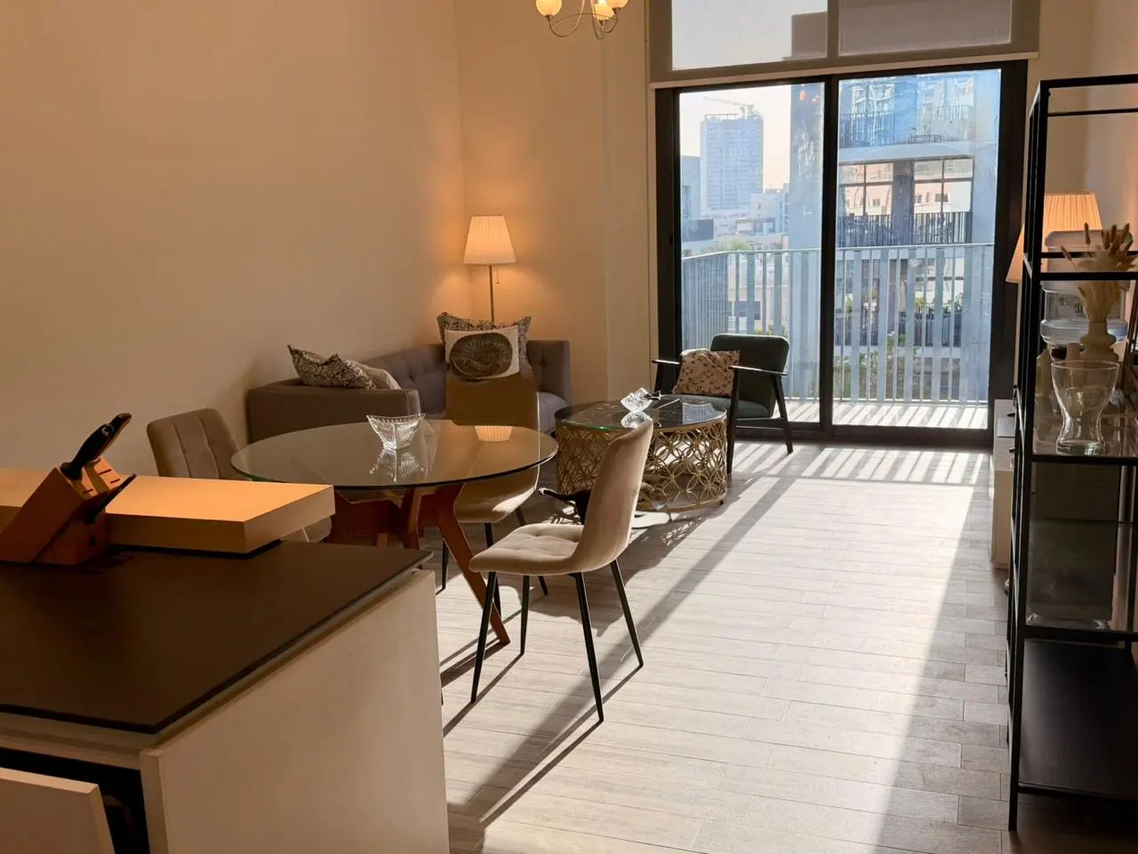 Sunlit apartment living room with a glass table, beige chairs, a gray sofa, and a balcony overlooking city buildings.