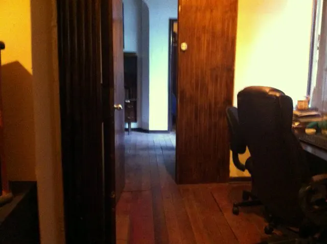 A view of a room with hardwood floors, a dark office chair, and an open wooden door leading to a brightly lit hallway.