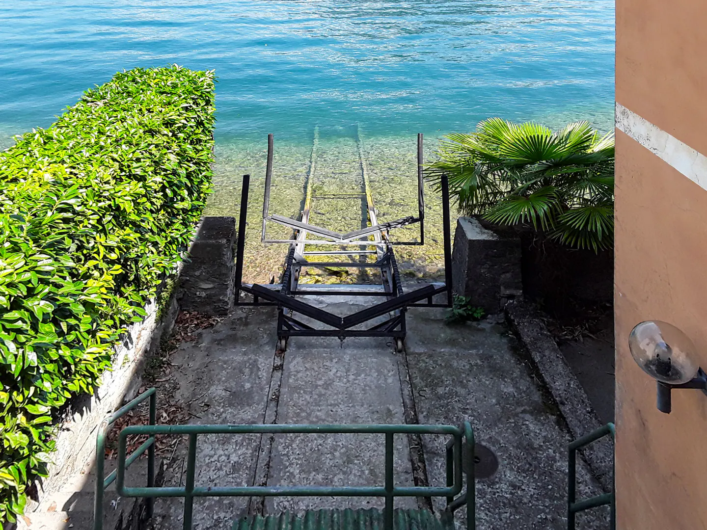 Concrete boat ramp leading into clear blue water, flanked by green foliage and a peach-colored wall with a light fixture.
