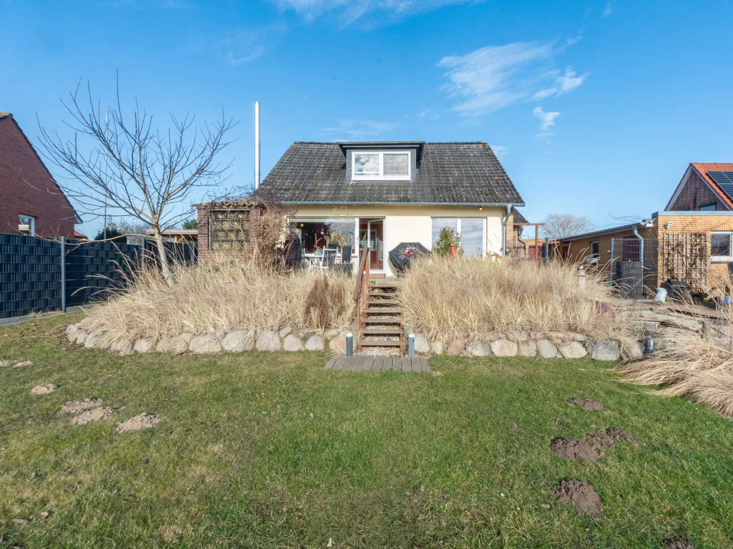 A single-story house with a gray roof, beige siding, and a green lawn under a blue sky. Tall, dry grass grows along a stone wall in front.