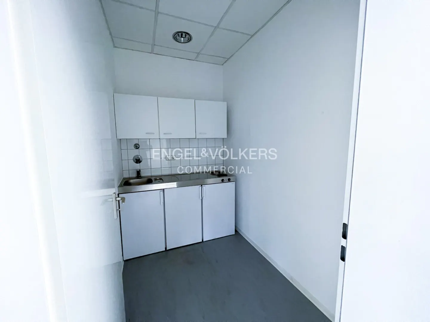 Small office kitchenette with white cabinets, stainless steel countertop, sink, and grey flooring in a minimalist room.