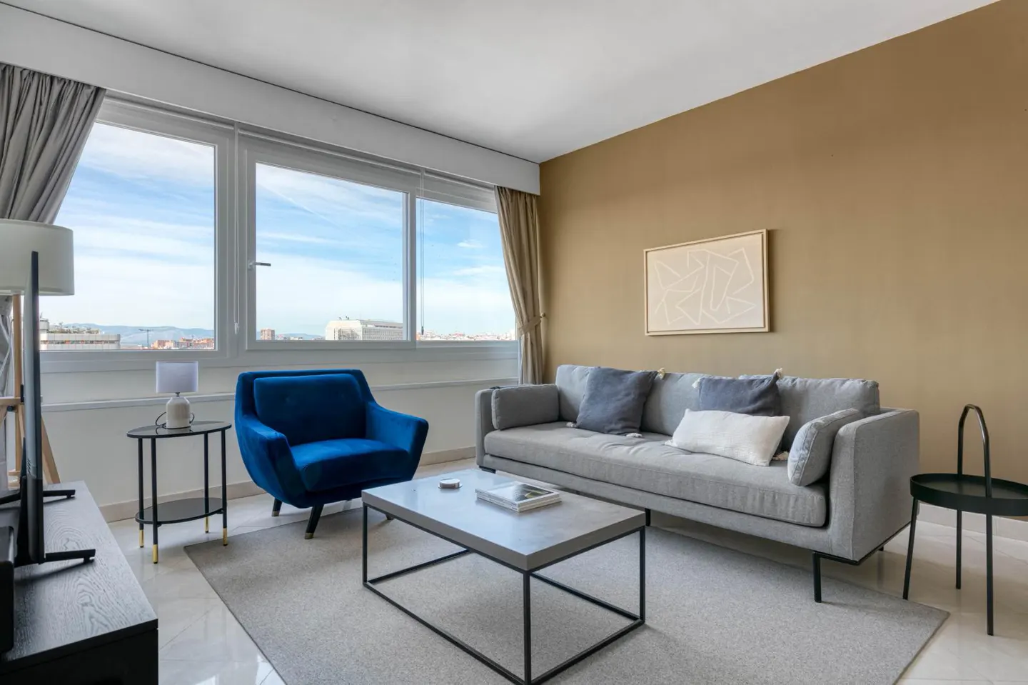 Living room with a gray sofa, blue armchair, and a concrete coffee table on a gray rug. Large windows offer a city view.