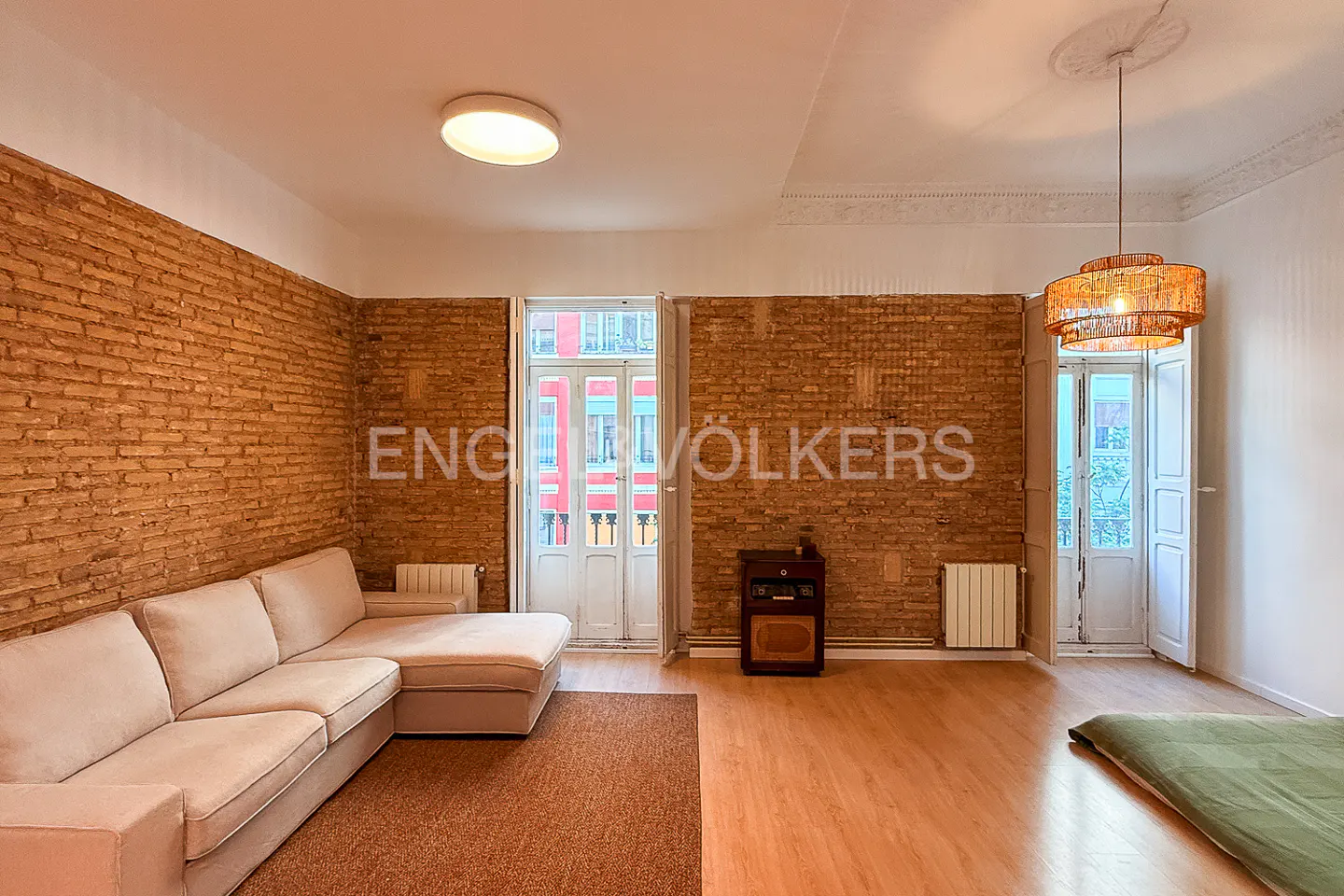 A living room with exposed brick walls, a beige sectional sofa, and a woven pendant light. White doors lead to a balcony.