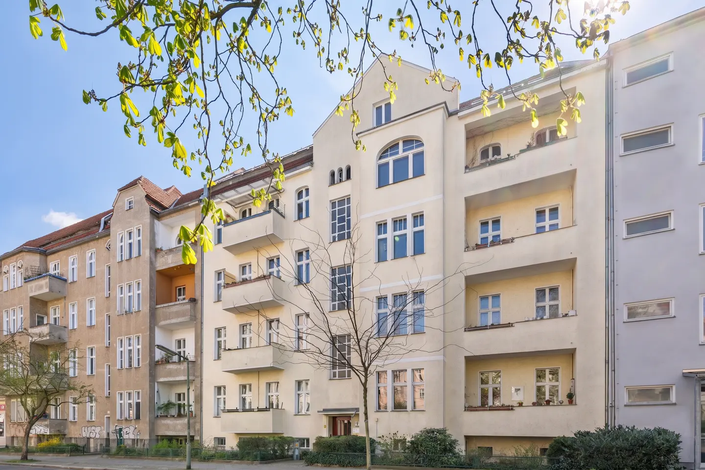 A cream-colored apartment building with balconies and large windows, framed by tree branches on a sunny day.
