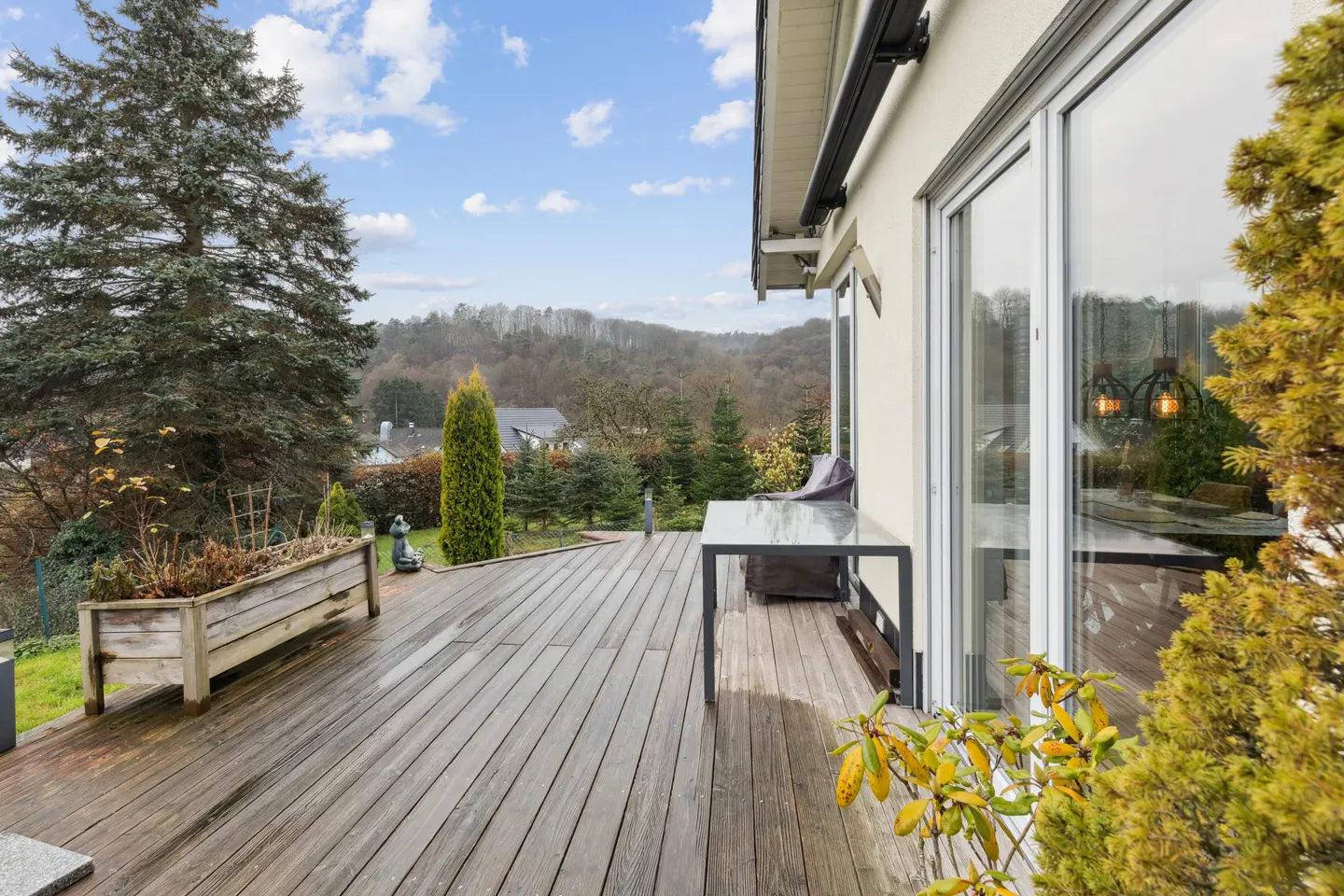 Exterior view of a wooden deck with a table and chairs, next to a house with large windows, overlooking a green landscape.