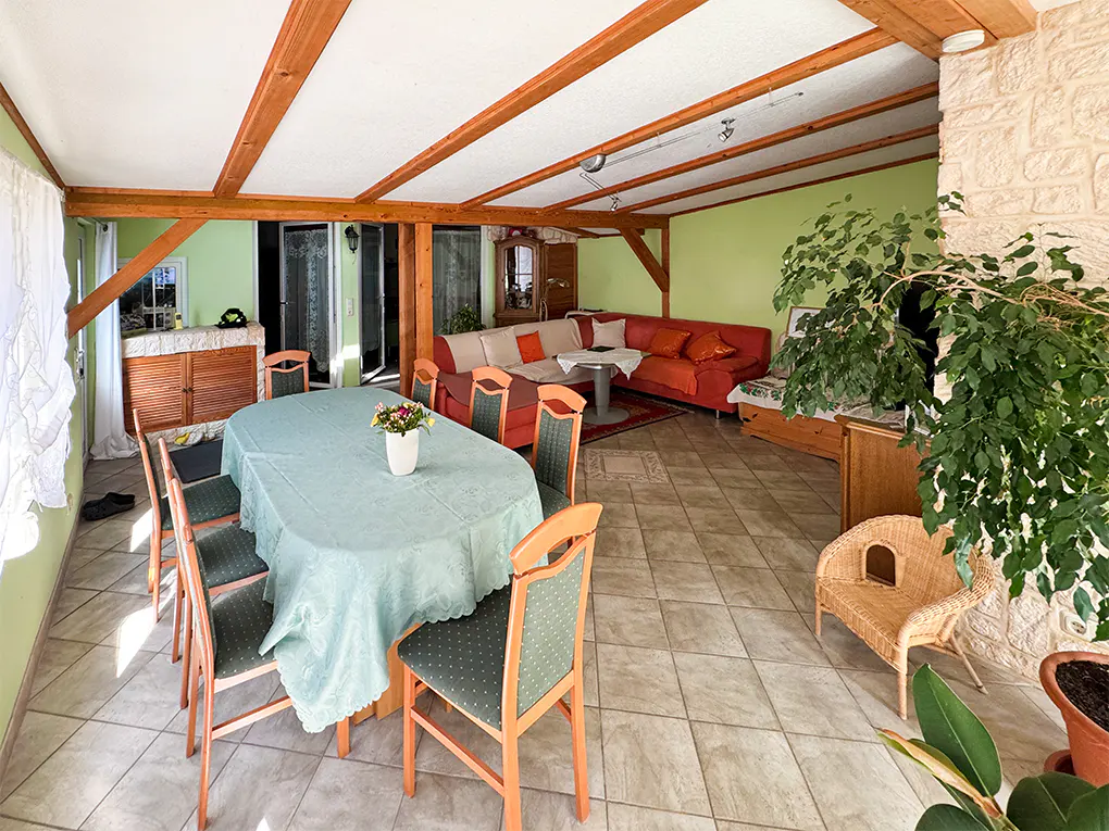 A sunroom with a dining table, red sectional sofa, and a large potted plant. The room has a tiled floor and wooden beams on the ceiling.