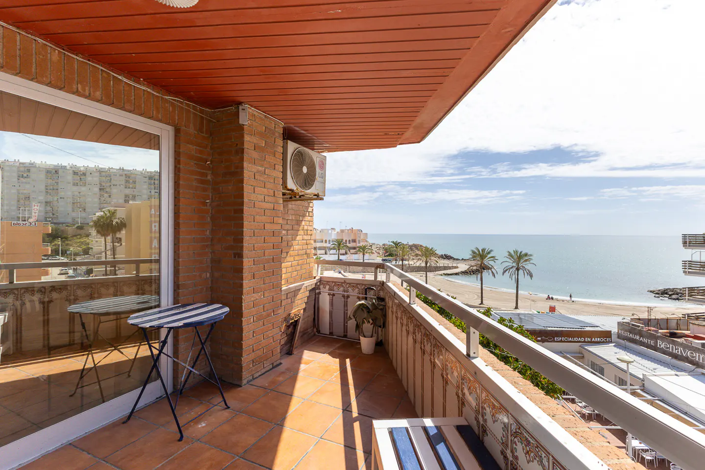 Balcony view of the beach with palm trees and blue sky. A small table sits near a sliding glass door.