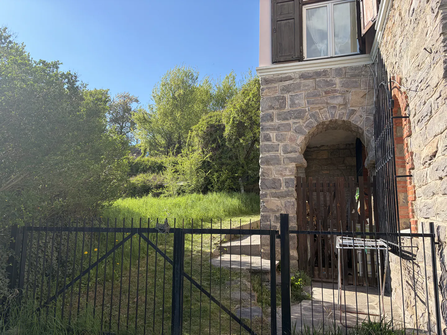 Exterior view of a stone house with an arched entrance, black iron fence, and green lawn.