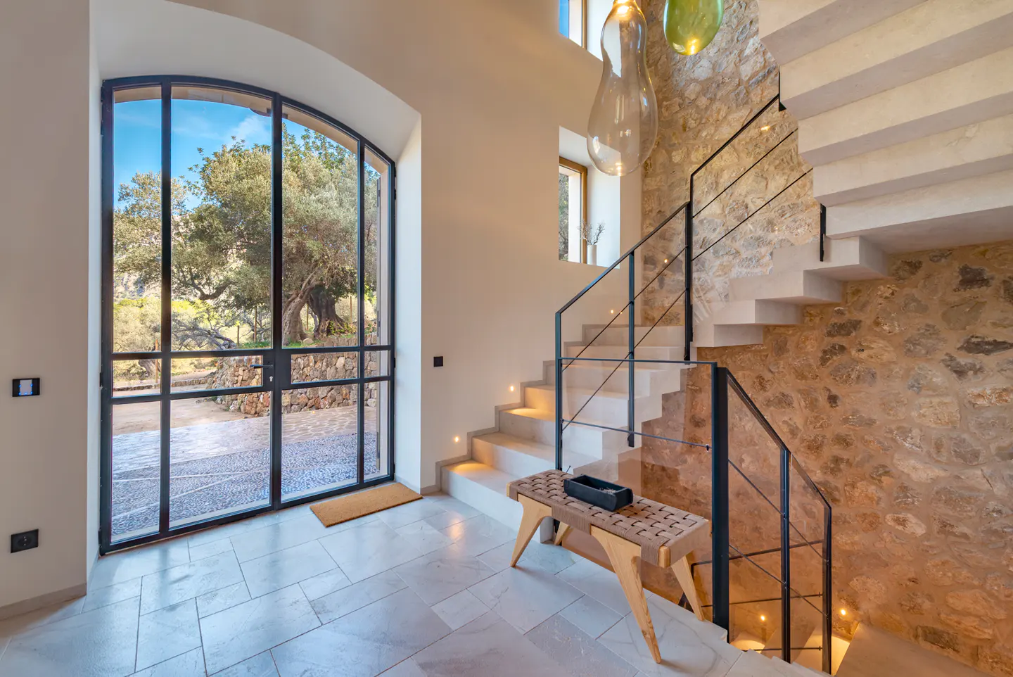 Interior view of a home's foyer with stone wall, staircase, bench, and large arched window looking out to trees.